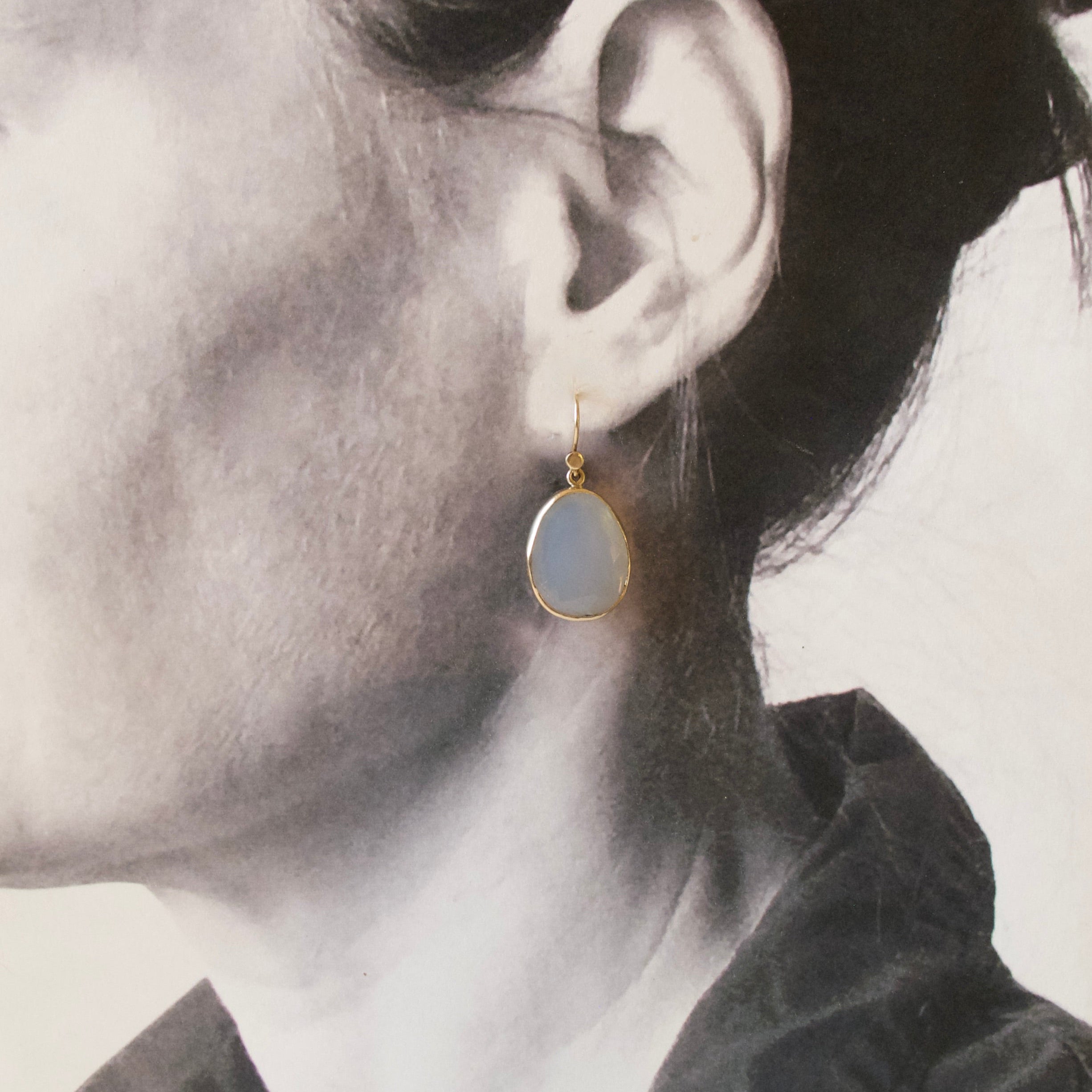 A close-up side profile of a woman wearing a small teardrop-shaped drop earring with a pale blue-gray stone and gold-tone setting.