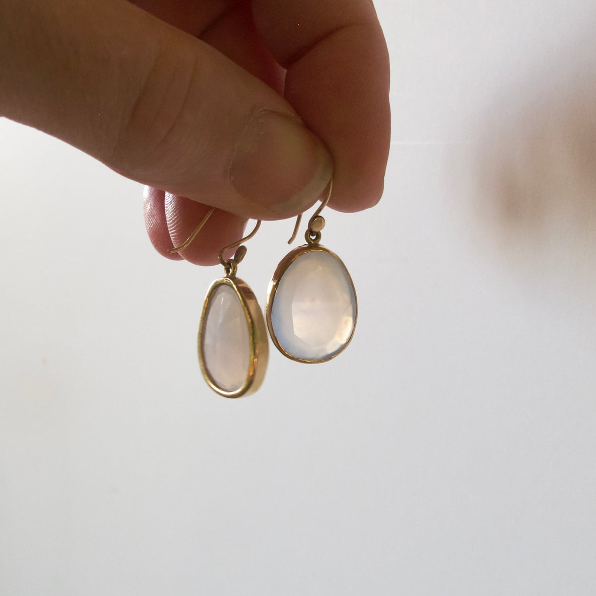 A close-up of a hand holding a pair of gold hoop earrings with oval milky white cabochon gemstones against a bright, soft-focus white background.