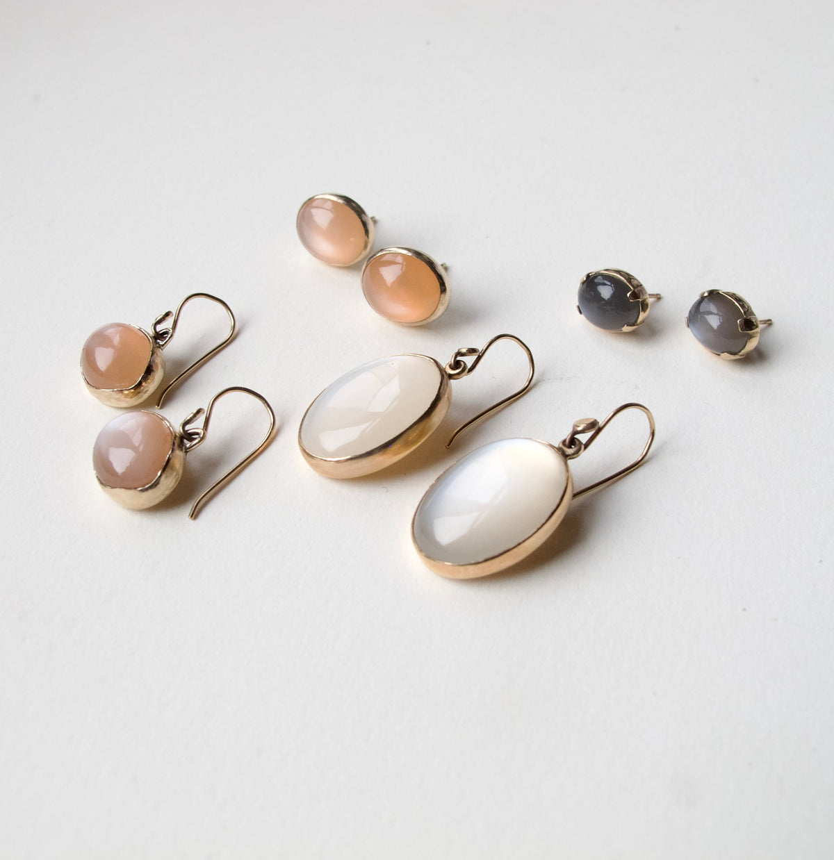 A set of gold-tone earrings and stud earrings arranged on a light off-white background. The collection includes three pairs of drop earrings: two with peachy-pink cabochon stones and one with a milky white cabochon stone, plus two pairs of stud earrings with gray cabochon stones.