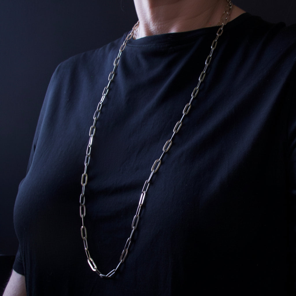 A close-up of a person wearing a long, chunky silver-toned paperclip chain necklace over a black t-shirt against a dark background.