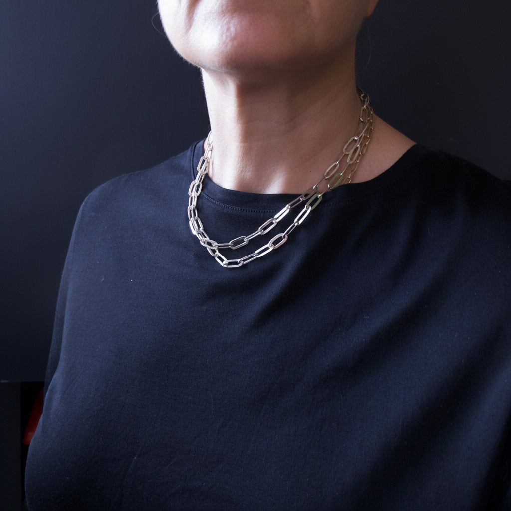 A close-up of a person wearing a layered chain necklace with elongated oval links over a black crew-neck shirt against a dark background.