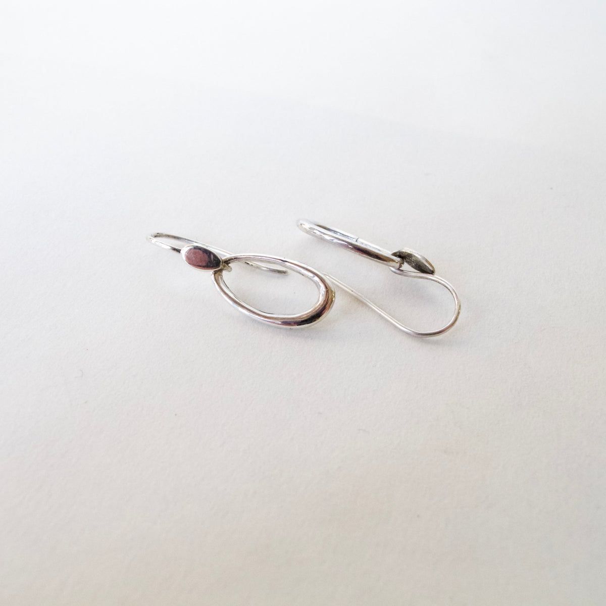 A pair of minimalist silver hoop earrings with leverback closures, displayed on a clean, light off-white background.