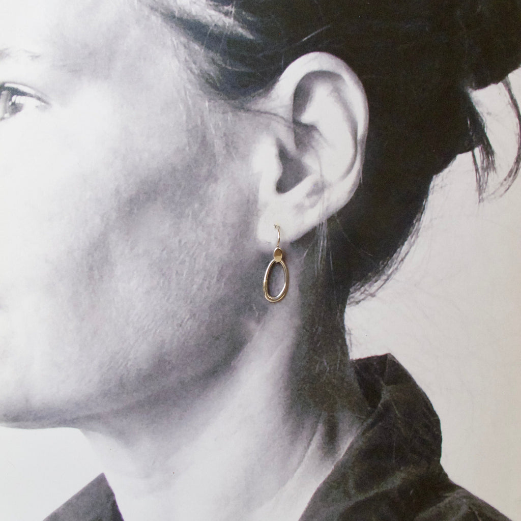 A close-up side profile of a woman wearing a small gold hoop earring with a dangling oval charm, shown against a light neutral background.