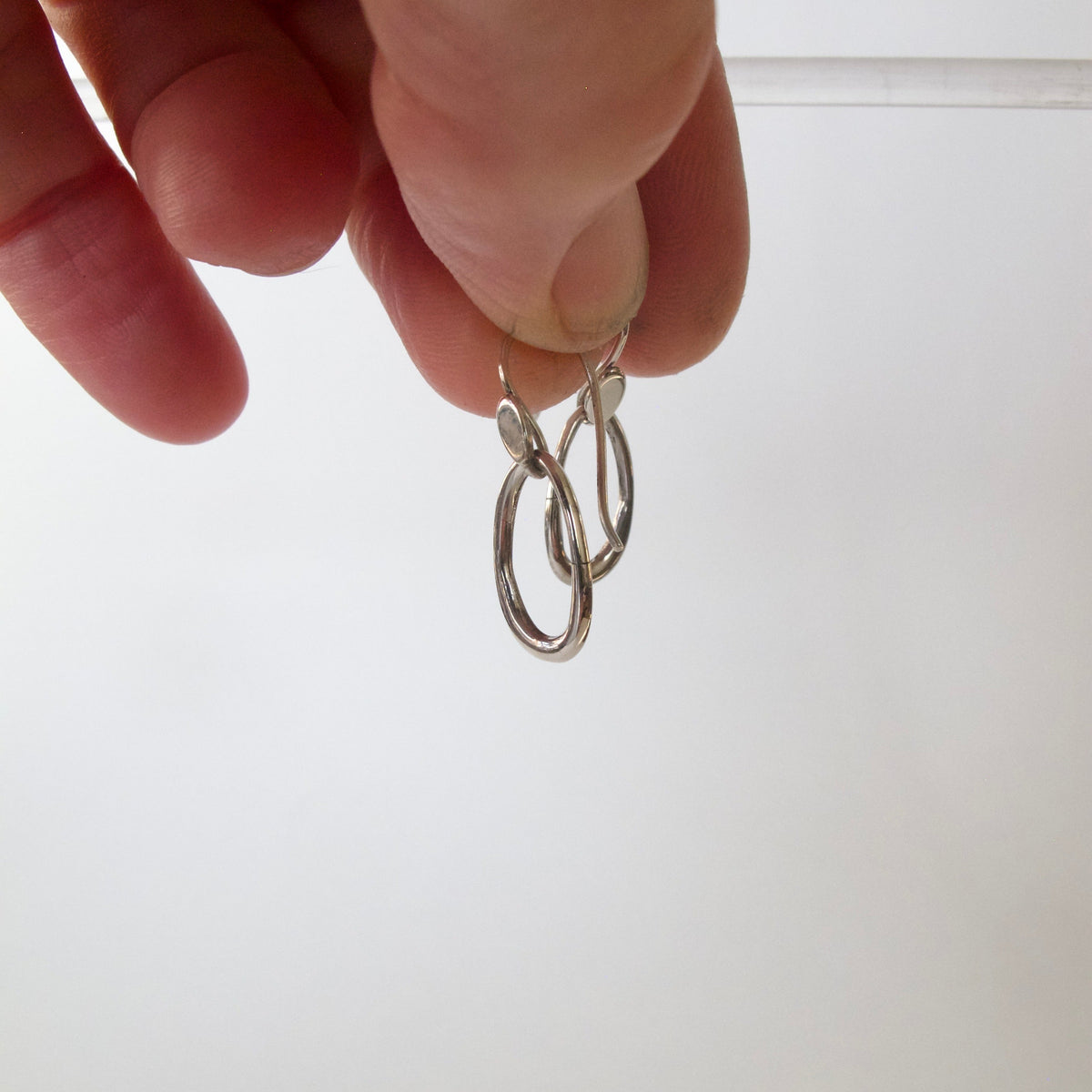 A close-up of a hand holding a pair of small silver hoop earrings against a plain light background.