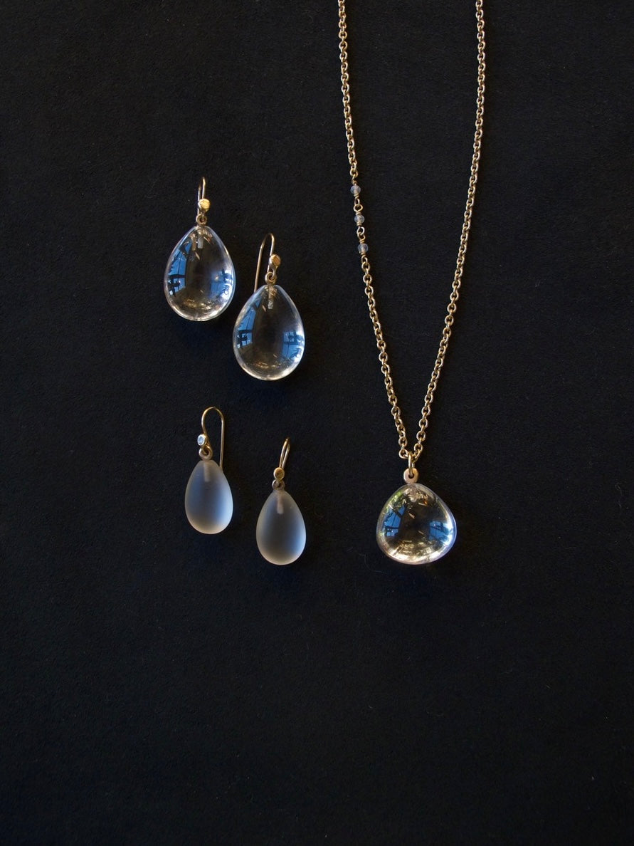 A jewelry set featuring a gold chain necklace and matching drop earrings with teardrop-shaped clear glass or acrylic pendants, displayed on a dark black background.