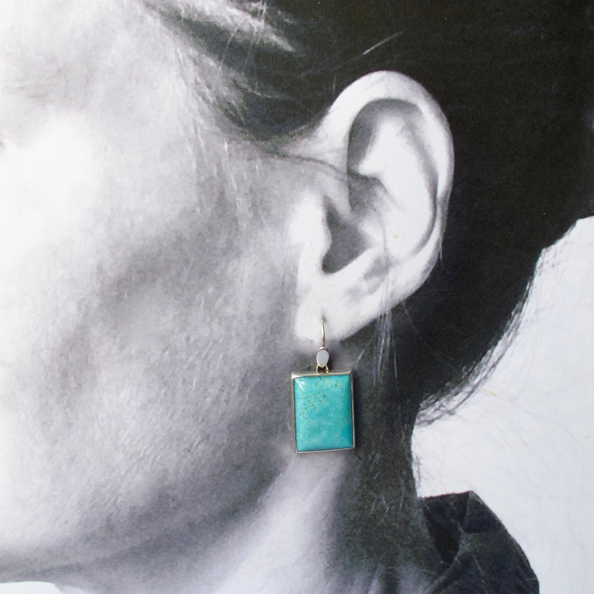 A close-up side profile of a person wearing a single square drop earring with a turquoise-blue stone in a silver-toned setting.
