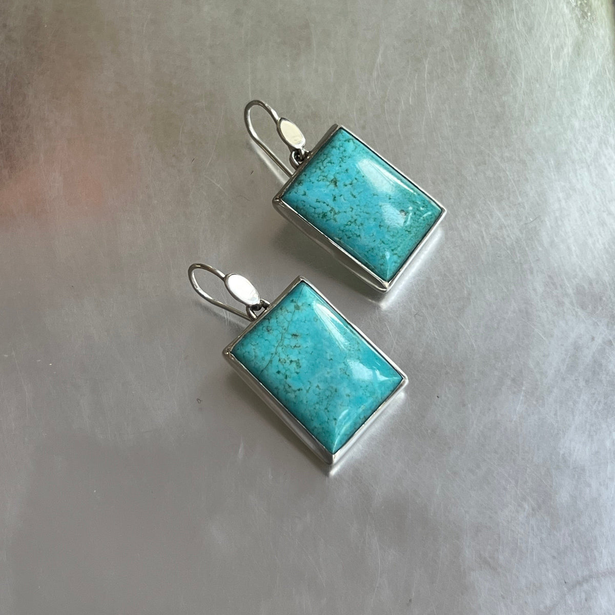 A pair of rectangular turquoise drop earrings with silver settings and fishhook ear wires, displayed on a textured gray surface.