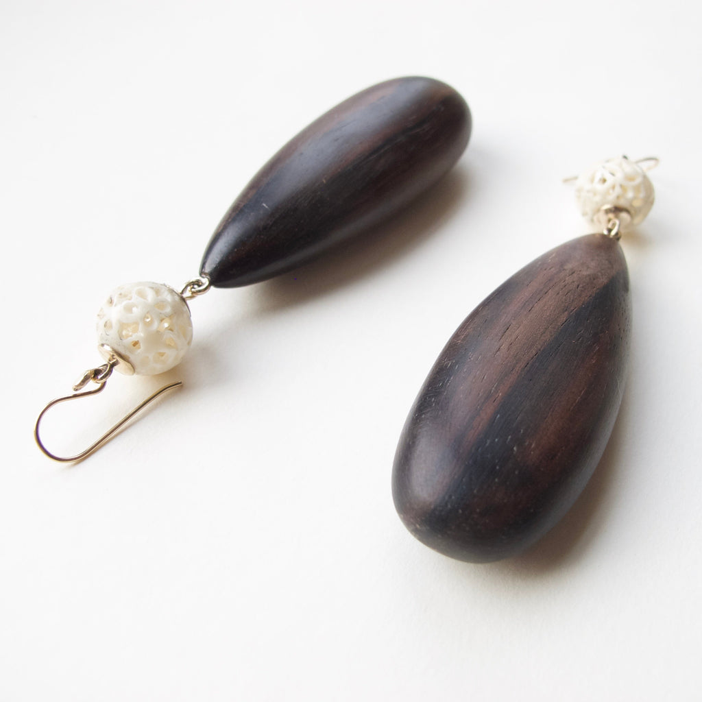 A pair of handmade teardrop dangle earrings made of dark brown wood, featuring a small white textured bead and a gold fishhook ear wire, displayed on a clean white background.