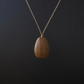 A teardrop-shaped wooden pendant necklace with a visible wood grain pattern, suspended from a thin gold chain against a dark black background.