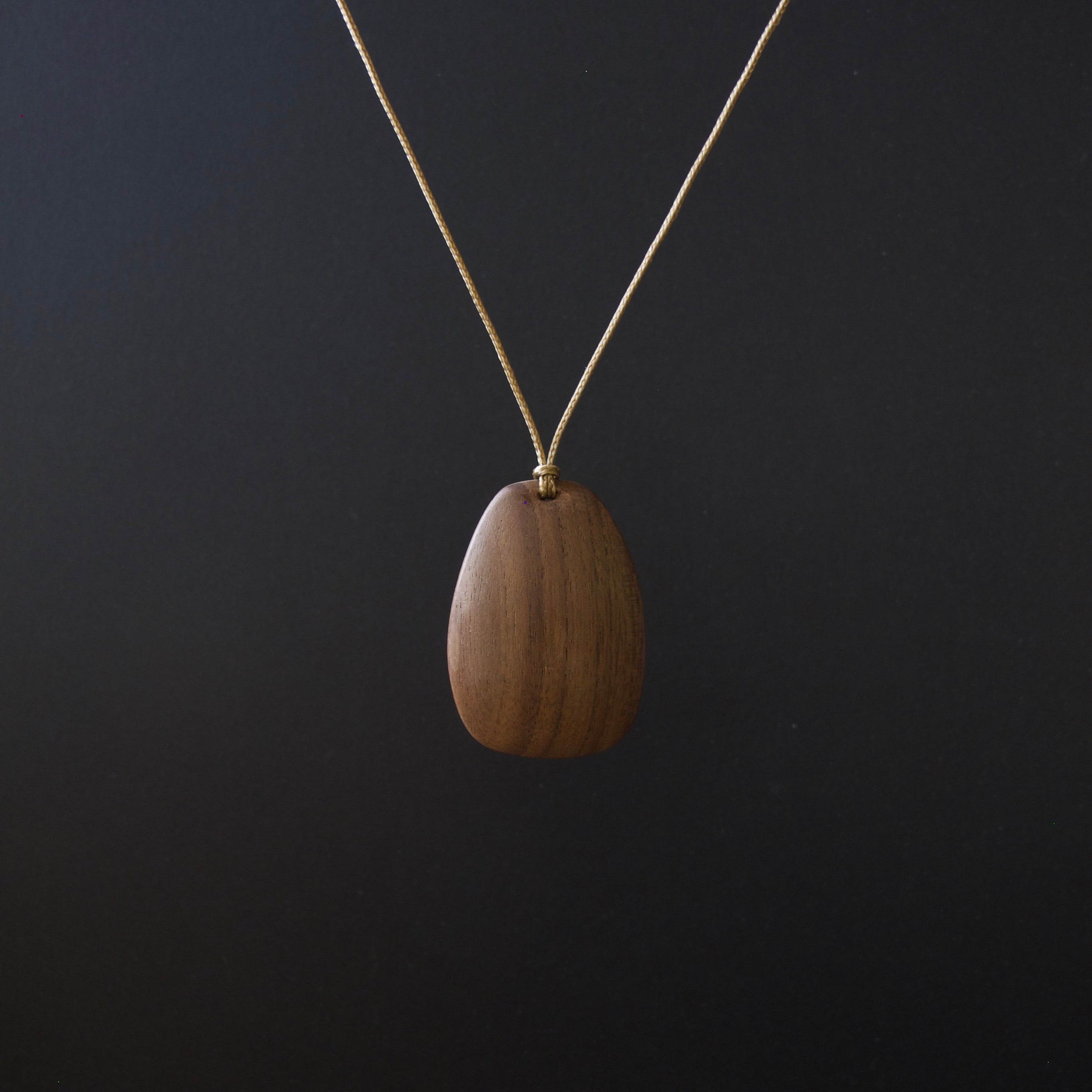 A teardrop-shaped wooden pendant necklace with a visible wood grain pattern, suspended from a thin gold chain against a dark black background.