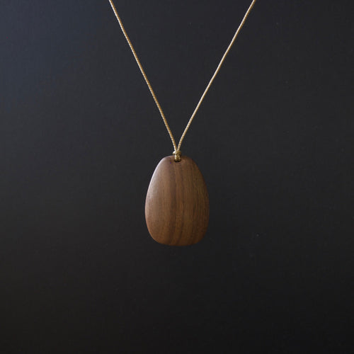 A teardrop-shaped wooden pendant necklace with a visible wood grain pattern, suspended from a thin gold chain against a dark black background.