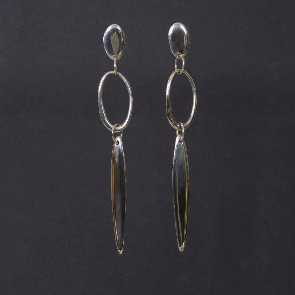 A pair of silver dangle earrings with a small oval stud, a larger open oval hoop, and a long teardrop-shaped drop, displayed against a dark gray background.