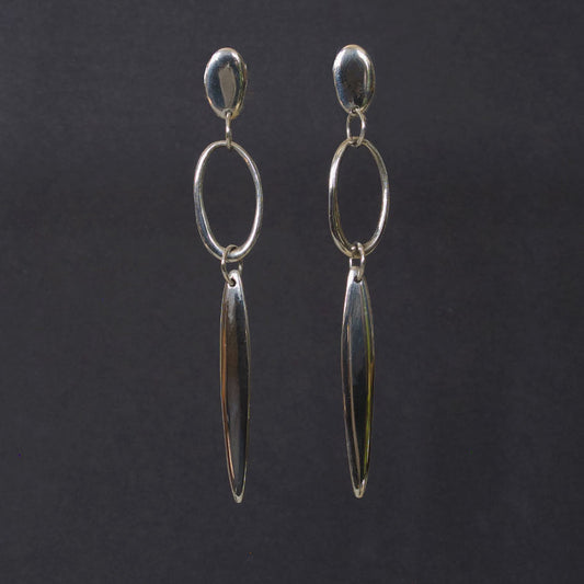 A pair of silver dangle earrings with a small oval stud, a larger open oval hoop, and a long teardrop-shaped drop, displayed against a dark gray background.