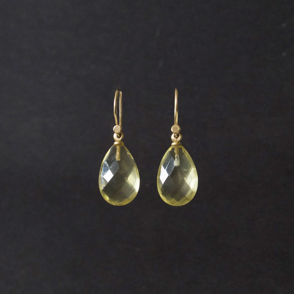 A pair of gold teardrop dangle earrings with faceted yellow-green gemstones, shown against a dark black background.