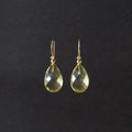 A pair of gold teardrop dangle earrings with faceted yellow-green gemstones, shown against a dark black background.