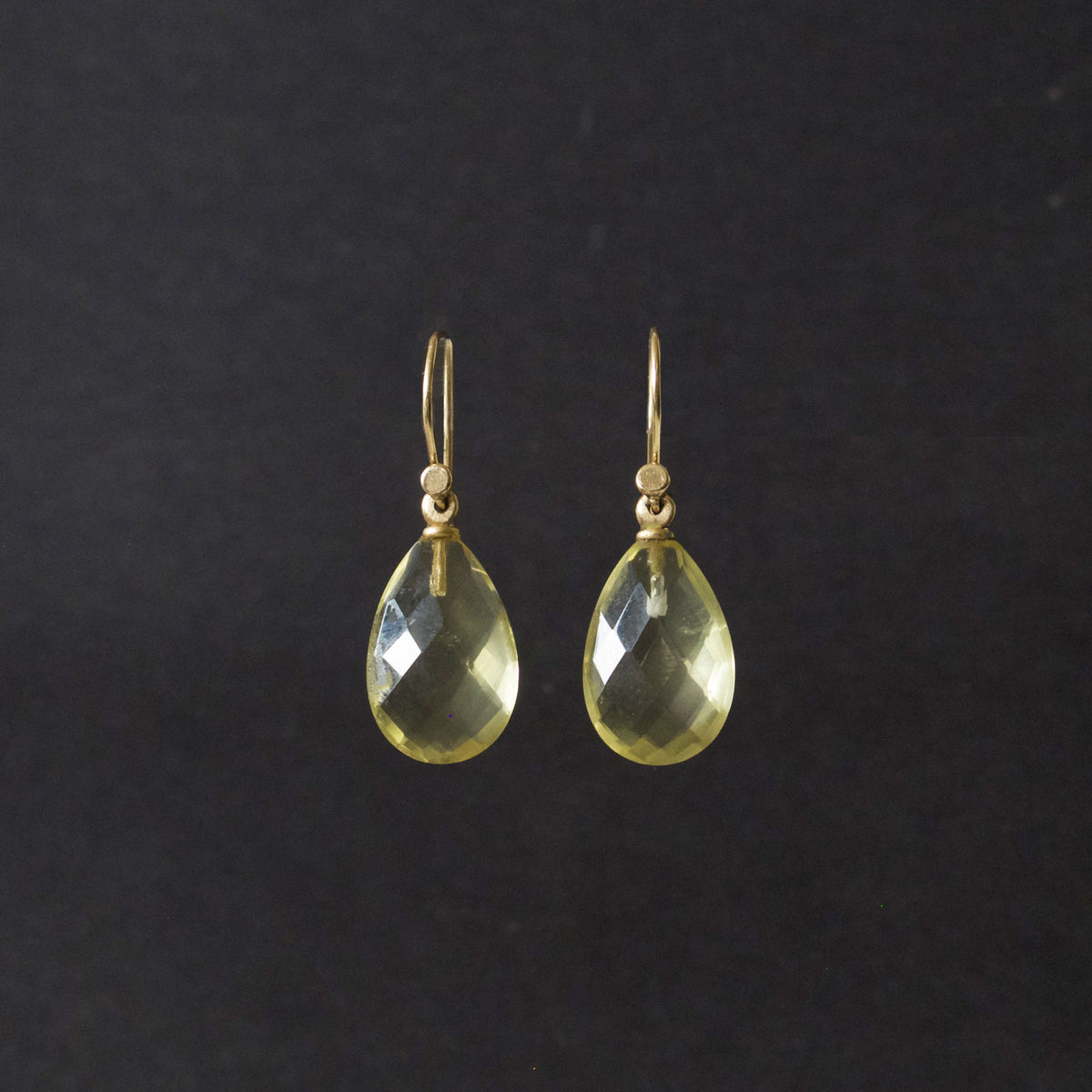 A pair of gold teardrop dangle earrings with faceted yellow-green gemstones, shown against a dark black background.