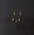 A pair of teardrop-shaped dark smoky glass drop earrings with gold hooks, displayed against a dark charcoal background.