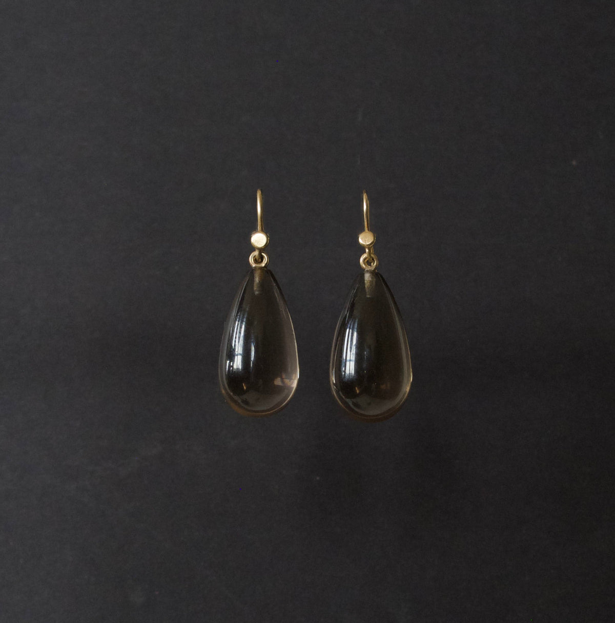 A pair of teardrop-shaped dark smoky glass drop earrings with gold hooks, displayed against a dark charcoal background.